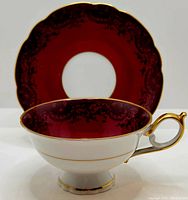 Front angled view of tea cup and saucer showing maroon interior and external white porcelain with gold trim and ornate dark patterns.