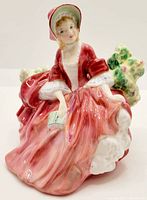 Front and side view of Royal Doulton Lydia figurine in red dress and bonnet, seated next to bush with flowers.