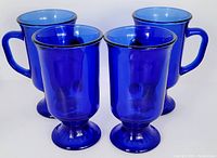 Four cobalt blue vintage glass mugs arranged together showing slight translucence and sturdy handles and footed bases.