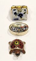 Three vintage trinket boxes shown: handbag shape with floral blue design, oval box with Arc de Triomphe scene, turtle-shaped cloisonné.