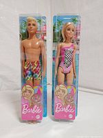 Photo showing Barbie and Ken dolls in original clear plastic packaging with tropical background, showcasing their swimsuits.
