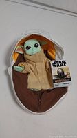 Front view of The Child plush secured in the plush pod, showing green face, tan robe, large black eyes, and attached tag with Star Wars Mandalorian branding.