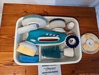 Top view of all components laid out in a white tray including handheld device, multiple brush attachments of varying texture and shape, cleaning solution bottle, and instruction manual.