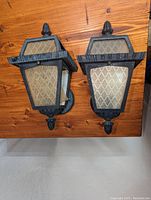 Pair of cast iron patio lights on wooden floor, showing front view and detailing of faded plastic panels and black metal frames.