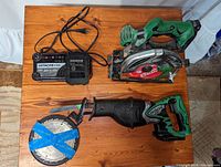 Photo of all items on wooden surface showing circular saw, reciprocating saw with blade, battery charger, battery, and extra circular saw blade taped with blue tape.