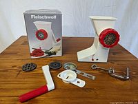 Photo showing Fleischwolf meat grinder attached to table with multiple accessories and original box in background.
