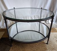 Full view of the round glass coffee table with metal frame and mirrored lower shelf on a carpeted floor.