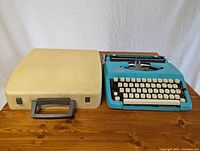 Blue K-Mart Deluxe 11 manual typewriter with white keys, placed on wooden table next to a white hard plastic carrying case with handle.