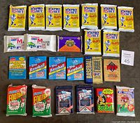 Top-down view of all 24 unopened sports wax packs arranged in rows on a dark surface showing a variety of mostly football packs with some baseball packs.