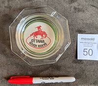 Top view of clear glass ashtray with red and black Ottawa Rough Riders logo and a red marker for scale.