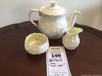 Three-piece Belleek tea set on a dark wooden table including the Serenity tea pot with lid and older sugar bowl and creamer side by side.