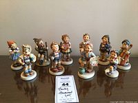 Lot of 10 Hummel porcelain figurines arranged in a semi circle, all standing on white circular bases, showing various activities and wearing traditional children's clothing.