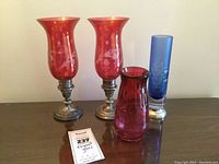 Pair of cranberry cut-to-clear hurricane shades on sterling silver bases, blue cylinder vase and magenta vase on wood surface