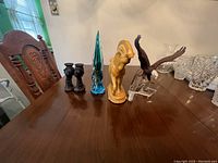 Photo shows multiple animal figurines including blue glass bird, yellow ceramic horse, glass or ceramic bald eagle on clear base arranged on wooden table.