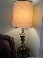 Side view of vintage gold tone metal lamp with beige fabric shade on a wooden side table next to a brown leather chair.