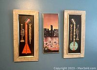 Set of three framed wall hangings on a white wall; two musical instrument themed pieces flank a cityscape with floral art piece.