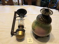 Front view of both antique oil lamps on a table, showing the caboose lamp with its round reflector and clear glass chimney and the floral-patterned round oil table lamp base.