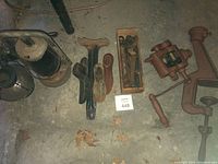 Photo of entire lot showing the shoe forms, lanterns, antique vice with pulley system, wooden box with tools, and box of nails on concrete floor.