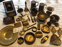 Photo showing extensive brass collection including ship's bell in box, tankards, bowls, ashtrays, utensils and various small brass decorative items spread on a flat surface.