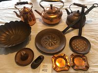 Photo showing three copper kettles with wooden handles, two copper bowls, a slotted spoon, two ashtrays, and a decorative shoe-shaped item on a table.