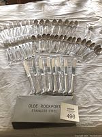 Full set of stainless steel flatware arranged on a surface with original box labeled Olde Rockport stainless steel visible. Pieces are individually wrapped and include knives, forks, spoons.