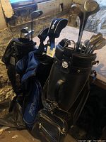 Three golf bags with assorted golf clubs inside, visible PING head covers and Callaway branding on one bag, placed together near a window.