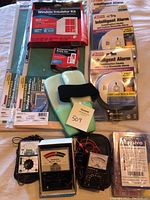 Photo showing two adjustable window screens by Frost King, window insulator kit, multiple Kidde combination smoke and carbon monoxide alarms, two analog multimeters, one digital multimeter, and a battery tester.