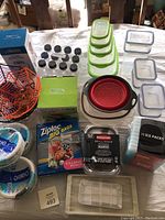 Wide top view of assorted plastic storage containers and other kitchen items on a table: green-lidded containers, black-capped bottles, Halloween basket, Ziploc bags, red-gray colander, Dixie bowls, ice packs.