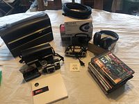 Photo showing vintage Kodak Instamatic camera with flash bulbs and black carrying case, 300 Series Polaroid case, large coil of coaxial cable, black wireless headphones, boxed Globe NOAA weather radio, and digital alarm clock on the table.