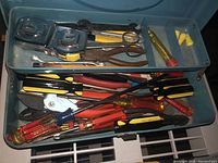 Top open view inside one metal toolbox showing various tools including tape measures, scissors, pliers, screwdrivers, and small parts like ear plugs.