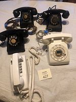 Five vintage telephones displayed on a flat surface, showing black and white rotary dials plus one push-button model.