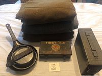 Stack of items including nested cast iron cooking utensils, metal first aid box, and cartridge box beside wool blankets.