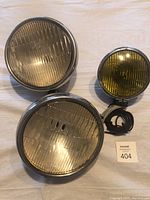 Three vintage headlamps displayed on a beige surface including two large round clear Ford headlamps and one smaller yellow lamp by Unity Manufacturing Co.