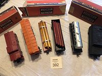 Six vintage Lionel model train cars arranged in a row with original boxes, showing diverse types including caboose, box cars, lumber flat beds with wooden loads, Texaco tanker, and tender.