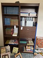 Two tall dark wood bookshelves filled with various books and organizers. The left shelf holds mostly blue and green hardcover books in sets along with vintage books and small paperback books on top shelf. The right shelf features white binders, stacked books, and books with varied covers on lower shelves.