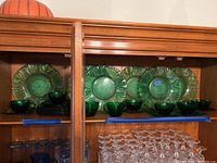 Display of six large 10.5-inch green glass plates together with six matching green glass cups and eight square saucers on a wooden shelf.