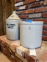 Two large beige stoneware pottery items; a jug marked Henry J. Barnett Albany NY and a #3 crock on a brick surface.