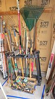 View of assorted garden tools including rakes with green plastic tines, shovels, axes, smaller hand tools in box, all held in a large black plastic tool stand.
