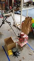 Full side view of Mantis tiller with long handles, red engine housing, and dirty bent tines. Replacement parts and small box are placed around the base.