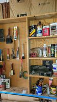 Wall-mounted pegboard with assorted paint brushes, some hanging, some in use showing wear, various paint and repair supplies on shelves beside it.
