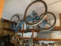 Vintage blue Schwinn Town and Country three-wheeler tricycle hanging upside down from garage rafters. Three wheels with metal fenders, rusted frame, visible wear and aged condition.