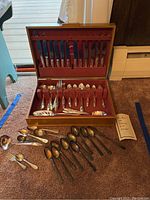 Full view of Oneida Tudor silverplate flatware set inside wooden chest including utensils and loose spoons scattered outside.
