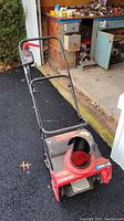 Front and handlebar view of the red and black Troy-Bilt snow thrower on driveway with garage in background.