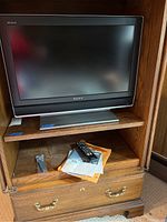 Sony Bravia 26 inch flat panel LCD HDTV shown inside a wooden cabinet with remote control and manual placed on the lower shelf inside the cabinet.