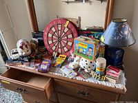 Wide view of dresser top showing entire lot including toys, games, lamp, piggy bank, lunchbox, and dartboard