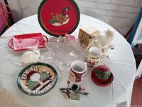 Front view of lot on round white table showing all items including Angel plate, mugs, red dish, metal tray and figurines.