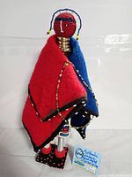 Front view of Ndebele doll showing red and blue fabric blanket, beaded head, and metal neck rings.