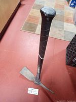 Full view of Husky pickaxe standing upright showing long black handle and metal pickaxe head with flat and pointed ends on the floor.