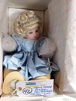 Porcelain doll seated in box wearing blue dress and silver tiara, with bubble wrap protecting hands and feet, tag attached with auction and doll details.