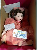 Porcelain doll inside box wearing pink floral dress and sheer pink overlay, with Certificate of Authenticity visible behind the doll.